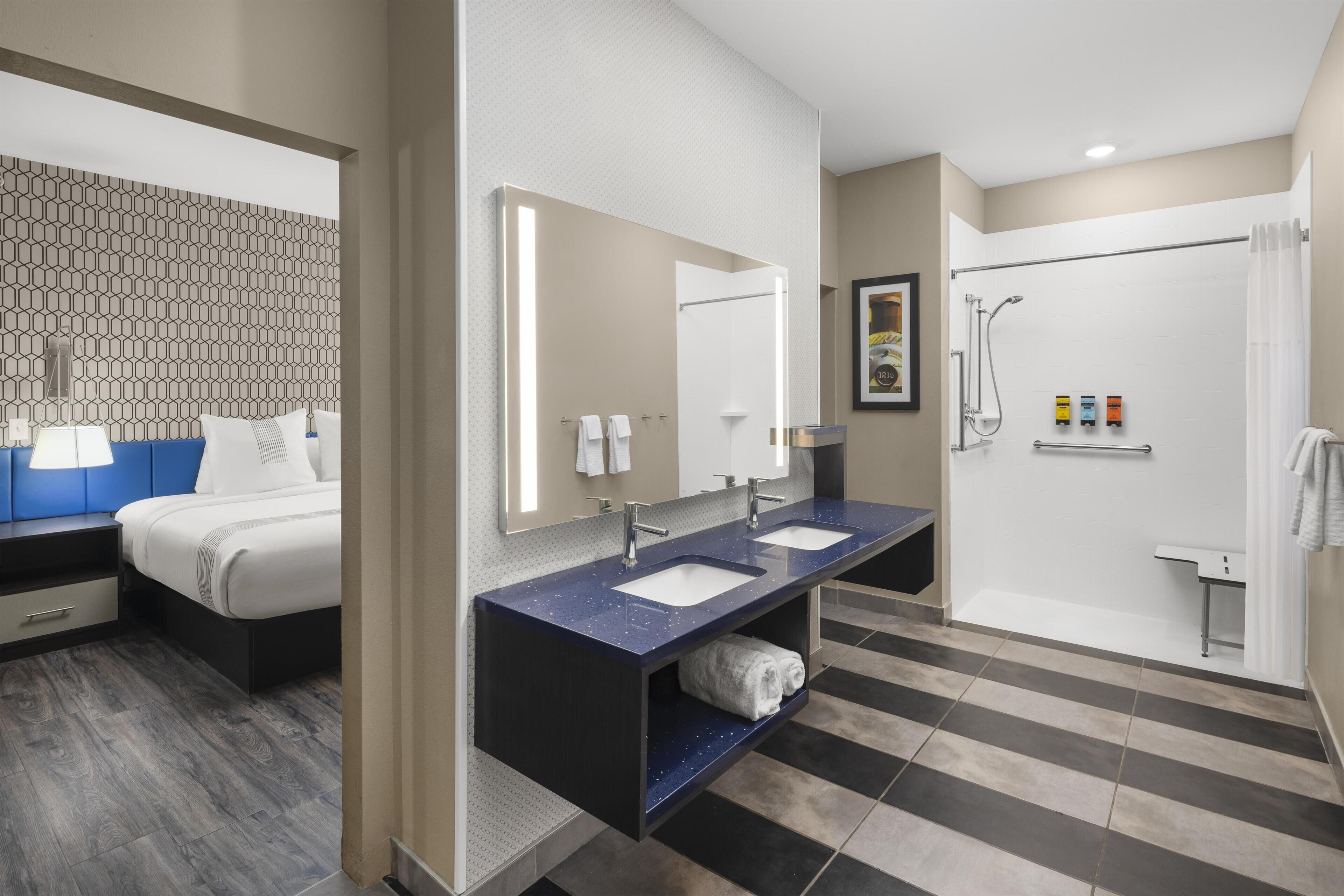 A modern hotel bathroom with a double sink vanity, a large mirror, a shower area, and a separate bedroom with a bed and nightstand.