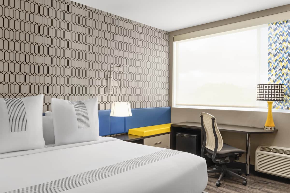 Habitación de hotel con una cama grande, un escritorio, una silla, una mesita, una lámpara y una ventana con cortinas estampadas.