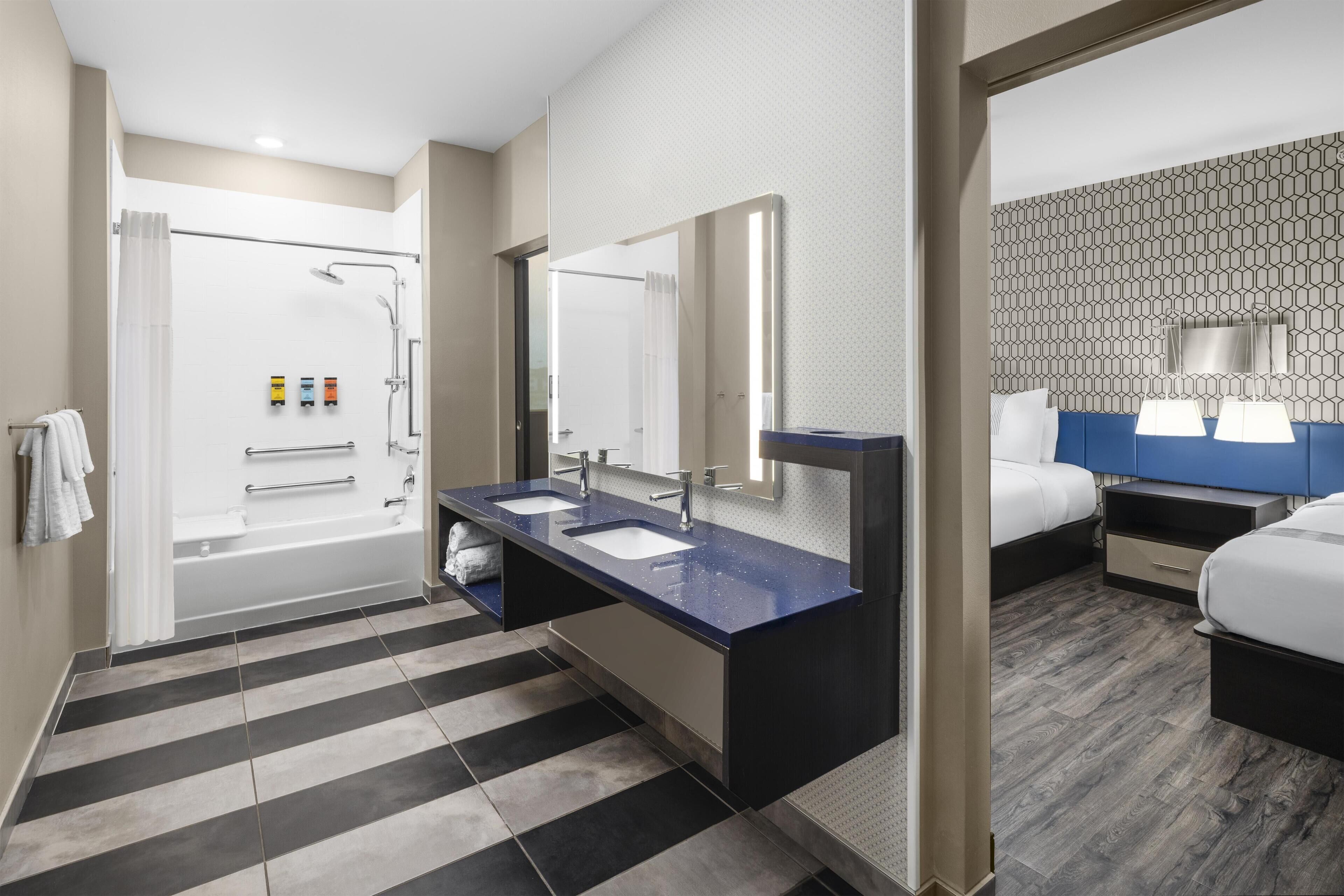 Ett modernt hotellbadrum med en stor spegel, två handfat, en dusch och ett separat sovområde med en säng och ett nattduksbord.