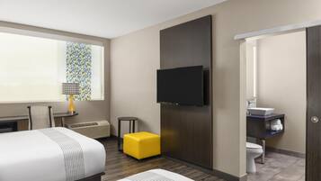 A hotel room with a bed, a TV mounted on the wall, a small table with a lamp, a yellow ottoman, and a bathroom with a sink and toilet.