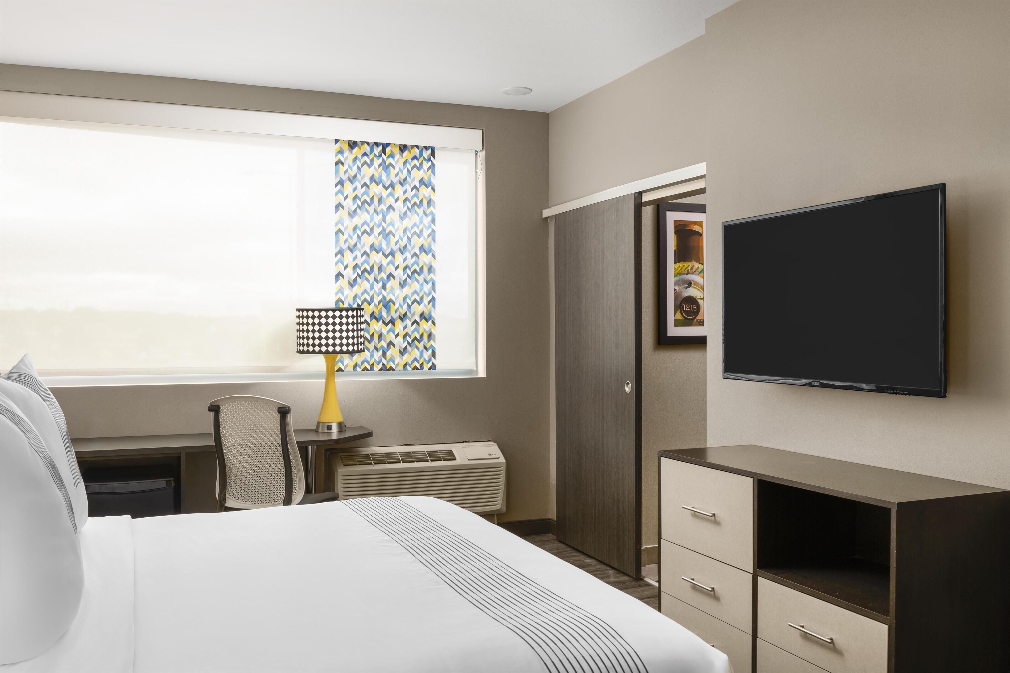 Ett hotellrum med en säng, ett skrivbord, en stol, en tv, en garderob och ett fönster med mönstrade gardiner.
