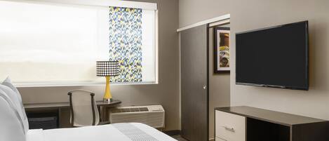 A hotel room with a bed, a desk, a chair, a television, a wardrobe, and a window with patterned curtains.