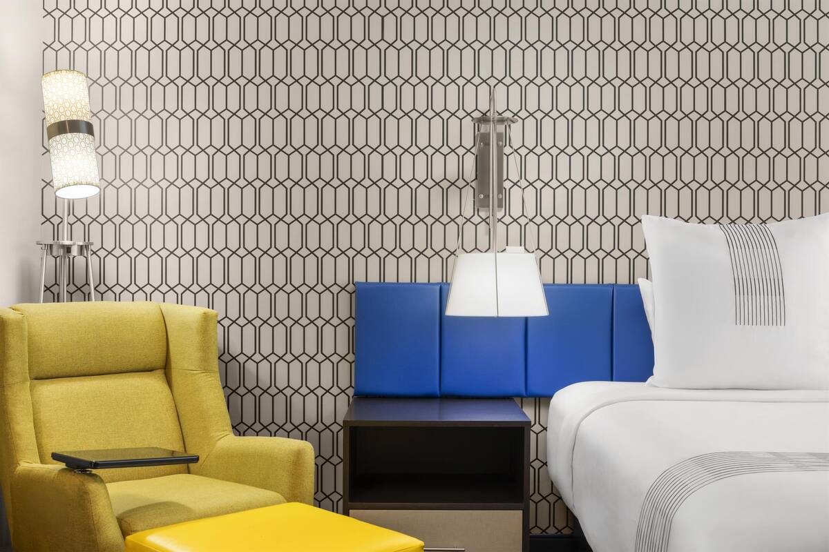 Hotelkamer met behang met patroon, een gele fauteuil, een blauw hoofdeinde, een nachtkastje met een lamp en een wit bed.