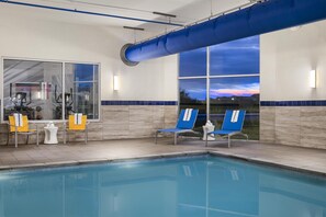 Zwembad met blauwe stoelen en fitnessapparatuur, een grote blauwe buis, en uitzicht op de ondergaande zon door grote ramen.