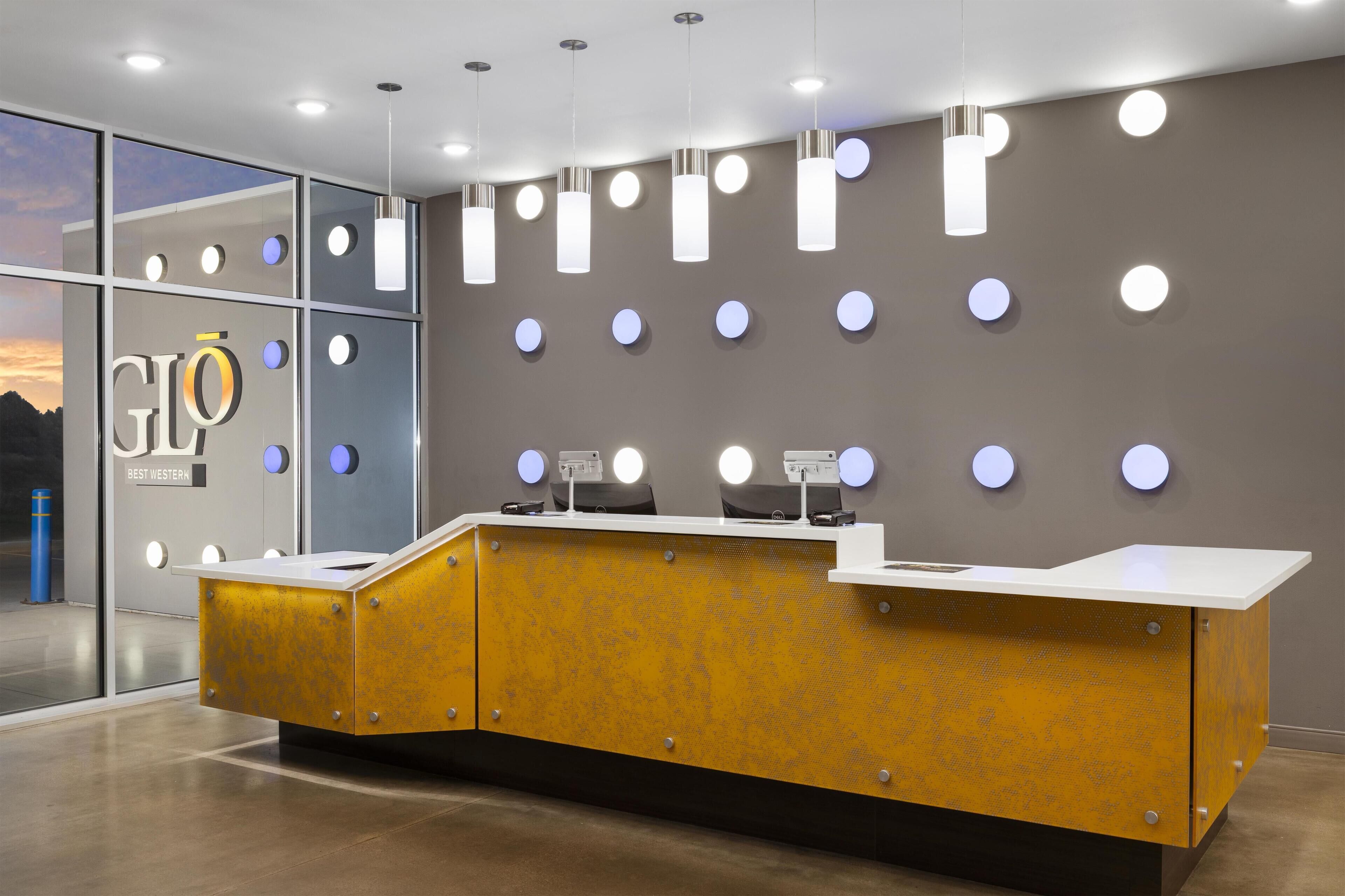 Un mostrador de recepción moderno con una combinación de colores amarillo y blanco, un muro de cristal con vistas a la ciudad y luminarias circulares en la pared.