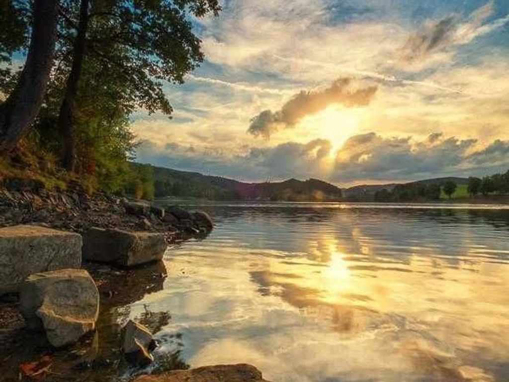 A calm lake with a sunset, trees, and a rocky shoreline.