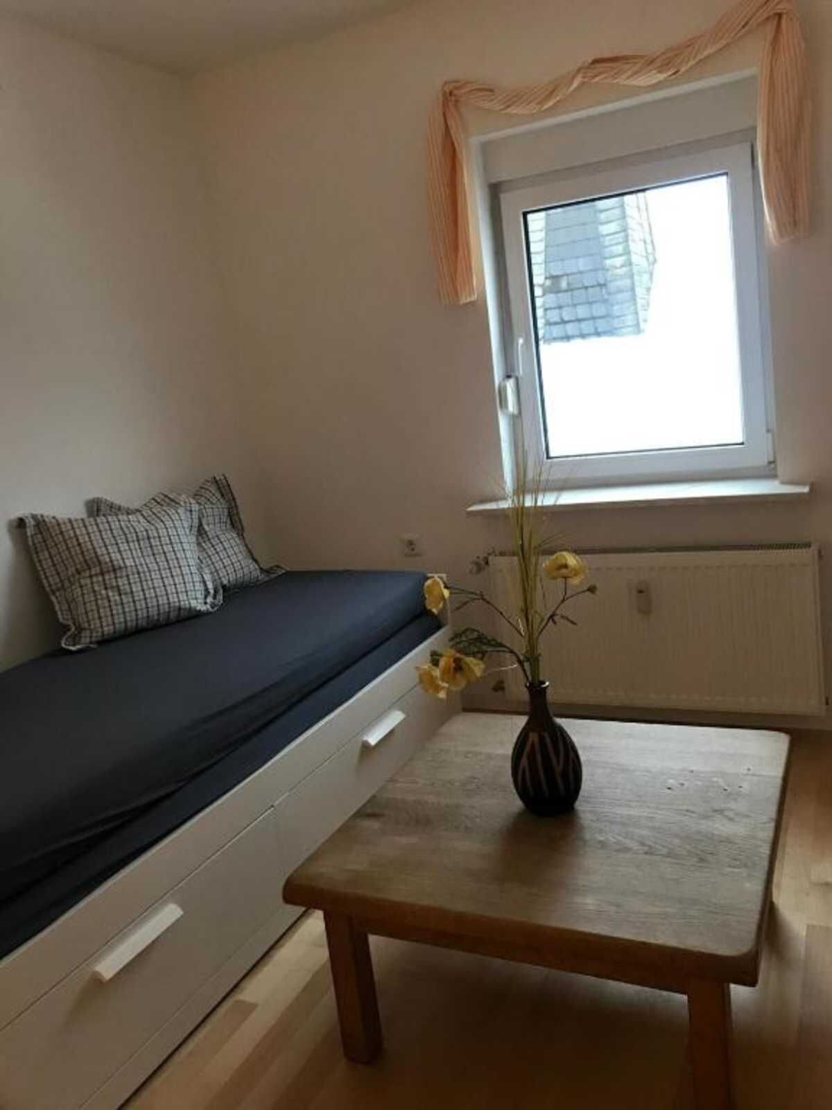 Une petite pièce avec un lit, une fenêtre avec des rideaux, une table avec un vase et un radiateur.