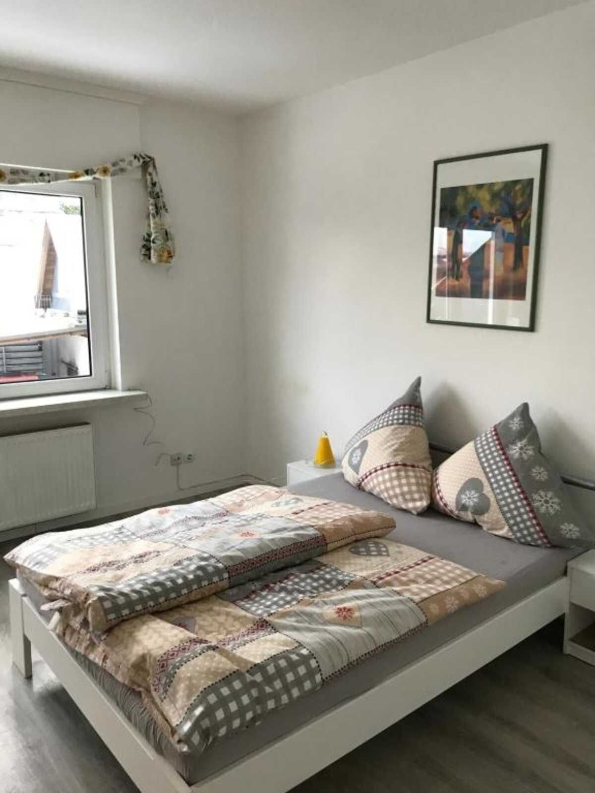 Ein modernes Schlafzimmer mit Bett, Fenster mit Blick, einem gerahmten Bild an der Wand und einem Nachttisch mit Lampe.