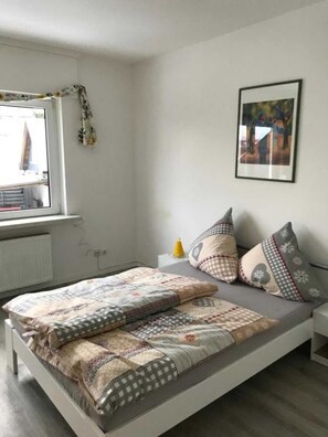 A modern bedroom with a bed, a window with a view, a framed picture on the wall, and a bedside table with a lamp.