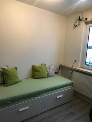 Een kleine eenpersoonskamer met een groene matras, twee kussens, een radiator en een raam.
