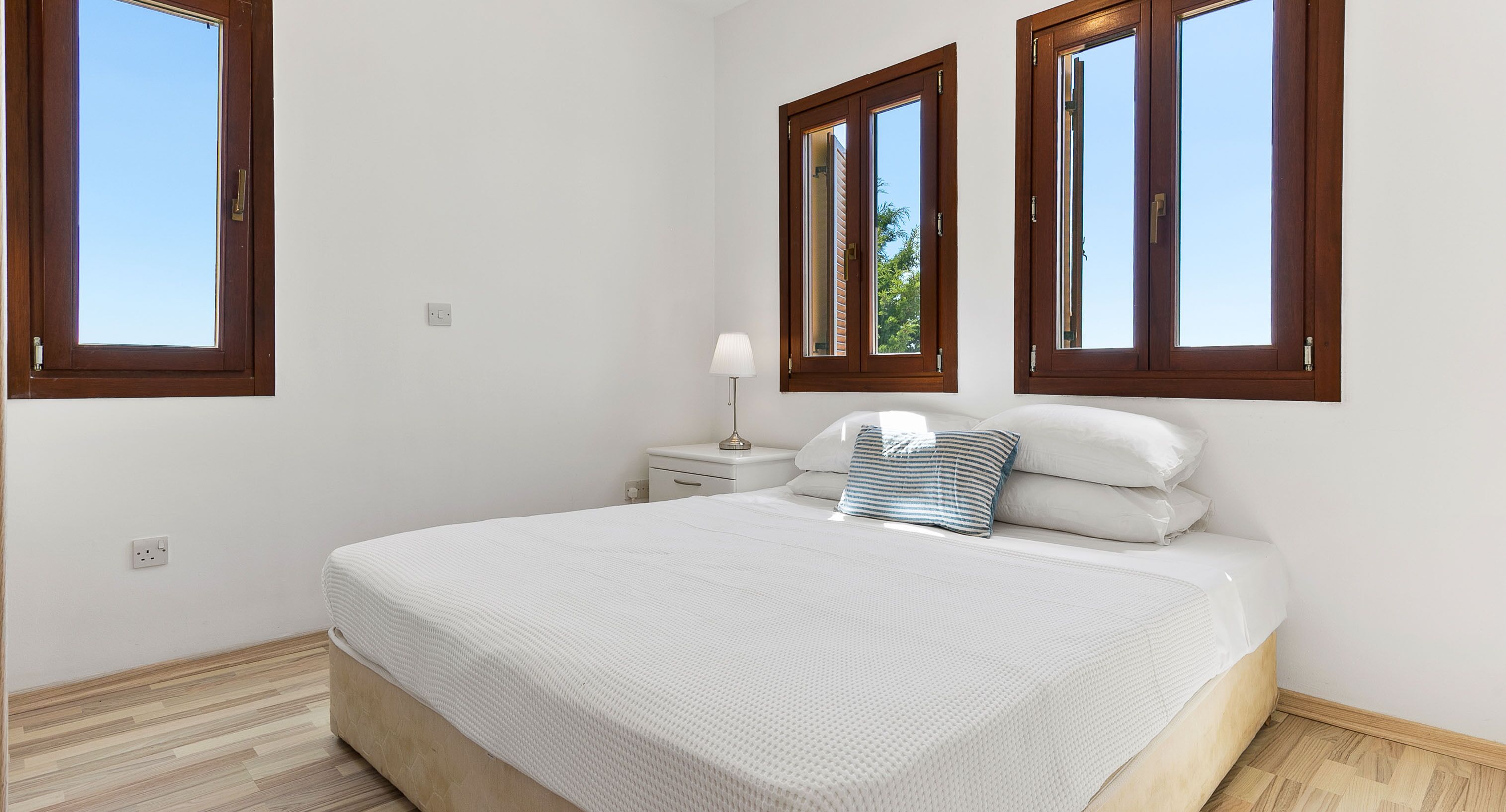 A bedroom with a large bed, a nightstand, a lamp, and three windows.