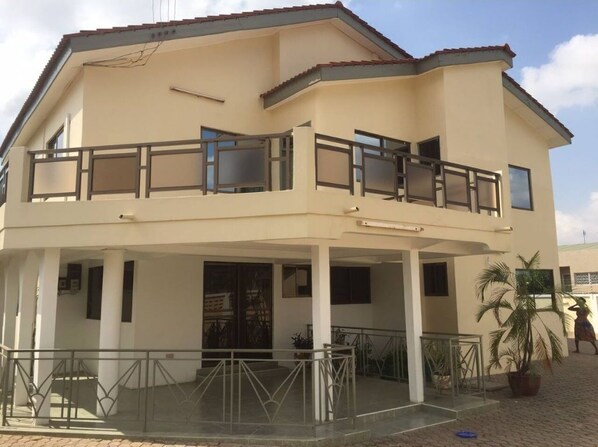 A two-story building with a balcony on the upper floor, a covered entrance, and a paved area with a palm tree.