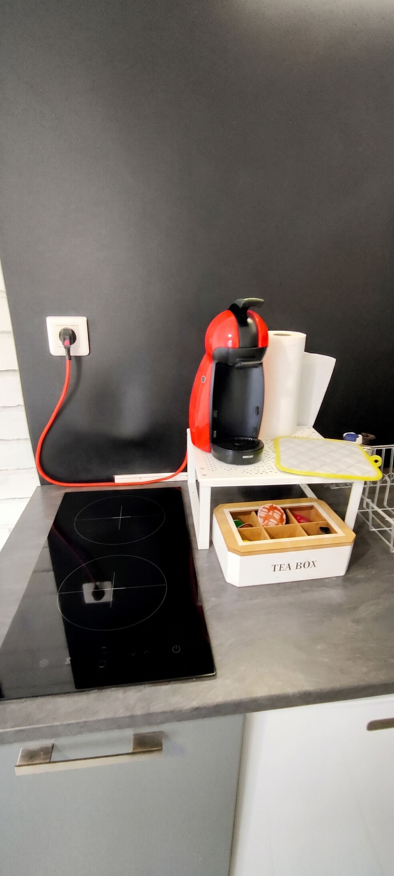 A kitchen with a black induction cooktop, a red coffee maker, a tea box, and a white kettle on a white countertop.