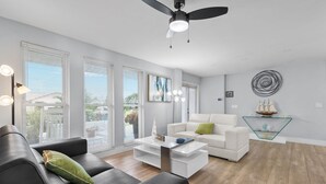 A modern living room with a white sofa, a black leather couch, a glass coffee table, and a ceiling fan.