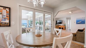 Un coin repas avec une table ronde et des chaises blanches, un téléviseur dans le salon et une vue sur l’extérieur grâce à de grandes portes vitrées.