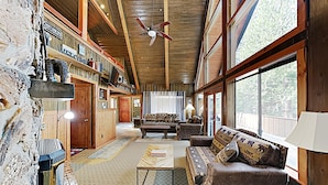 A spacious living room with a high ceiling, wooden beams, a fireplace, a sofa set, and a large window offering a view of the outdoors.