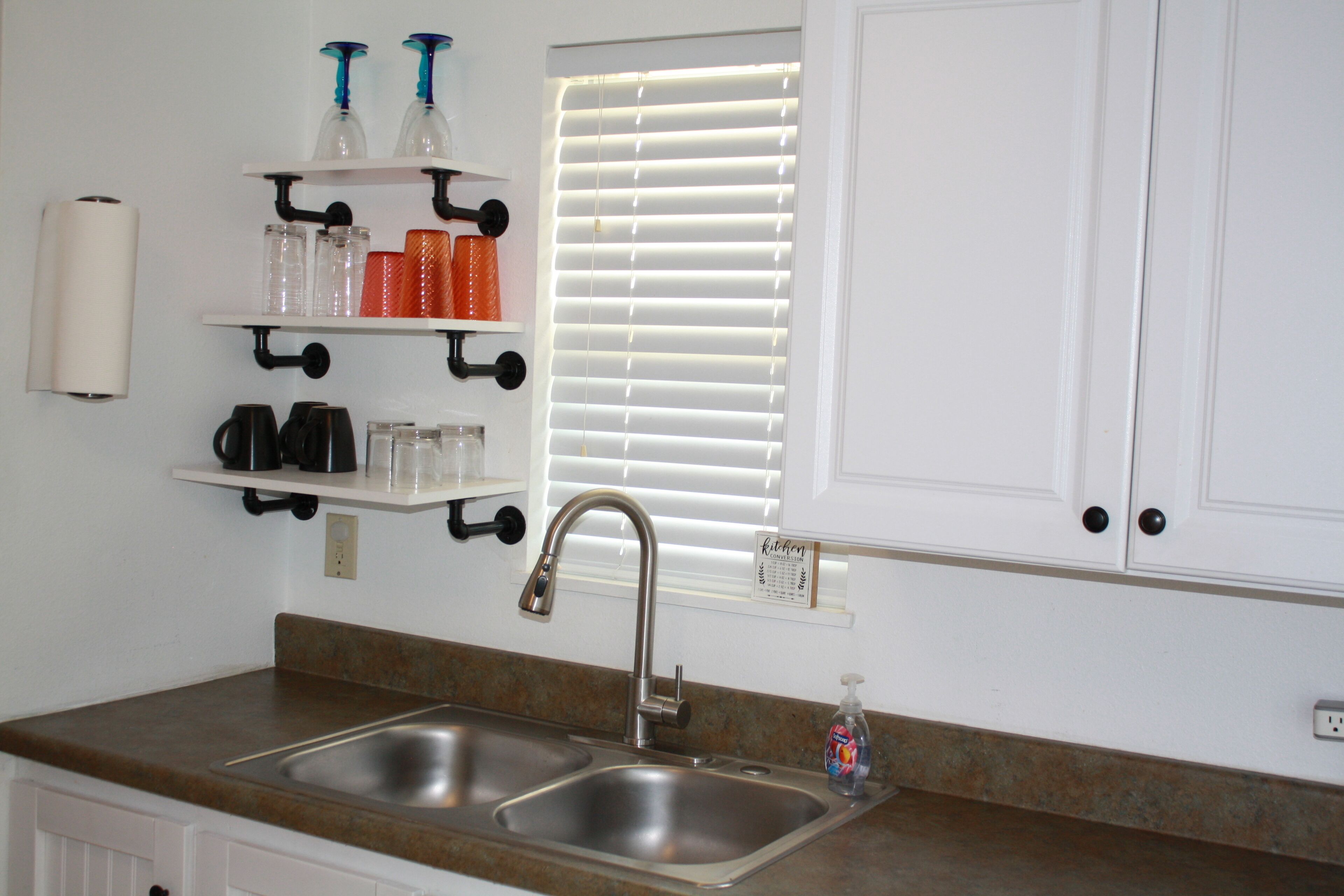 A kitchen with a double sink, stainless steel faucet, and a window with blinds. There are shelves above the sink holding various items.