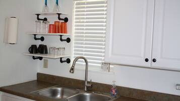 A kitchen with a double sink, stainless steel faucet, and a window with blinds. There are shelves above the sink holding various items.