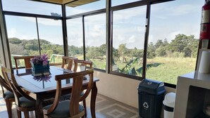 Een eetruimte met een tafel en stoelen, grote ramen met uitzicht op groen, een brandblusser en recyclingbakken.