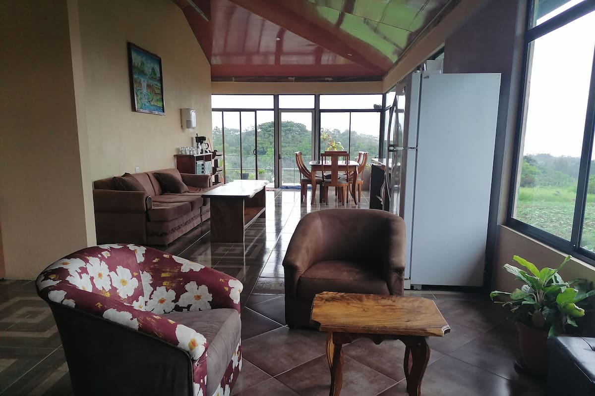 Ein großzügiger Wohnbereich mit Sofa, Sessel und Couchtisch. Durch große Fenster ist ein Essbereich mit Tisch und Stühlen zu sehen.