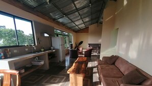 Een ruime woonkamer met een groot raam, een keukenhoek en een zithoek met een bruine bank en een houten salontafel.