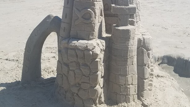 Un castell de sorra amb un teulat cònic i una base circular, situat a una platja sorrenca amb l'oceà al fons.