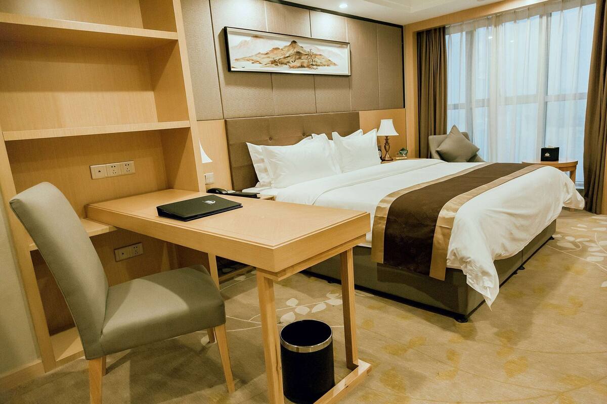 Une chambre d’hôtel avec un lit, un bureau, une chaise, une poubelle, une table de chevet, une lampe et un tableau au mur.