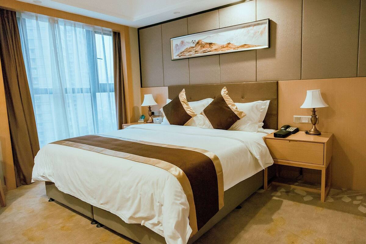 Une chambre d’hôtel avec un grand lit, deux tables de chevet, une commode, un tableau au mur et une fenêtre avec des rideaux.