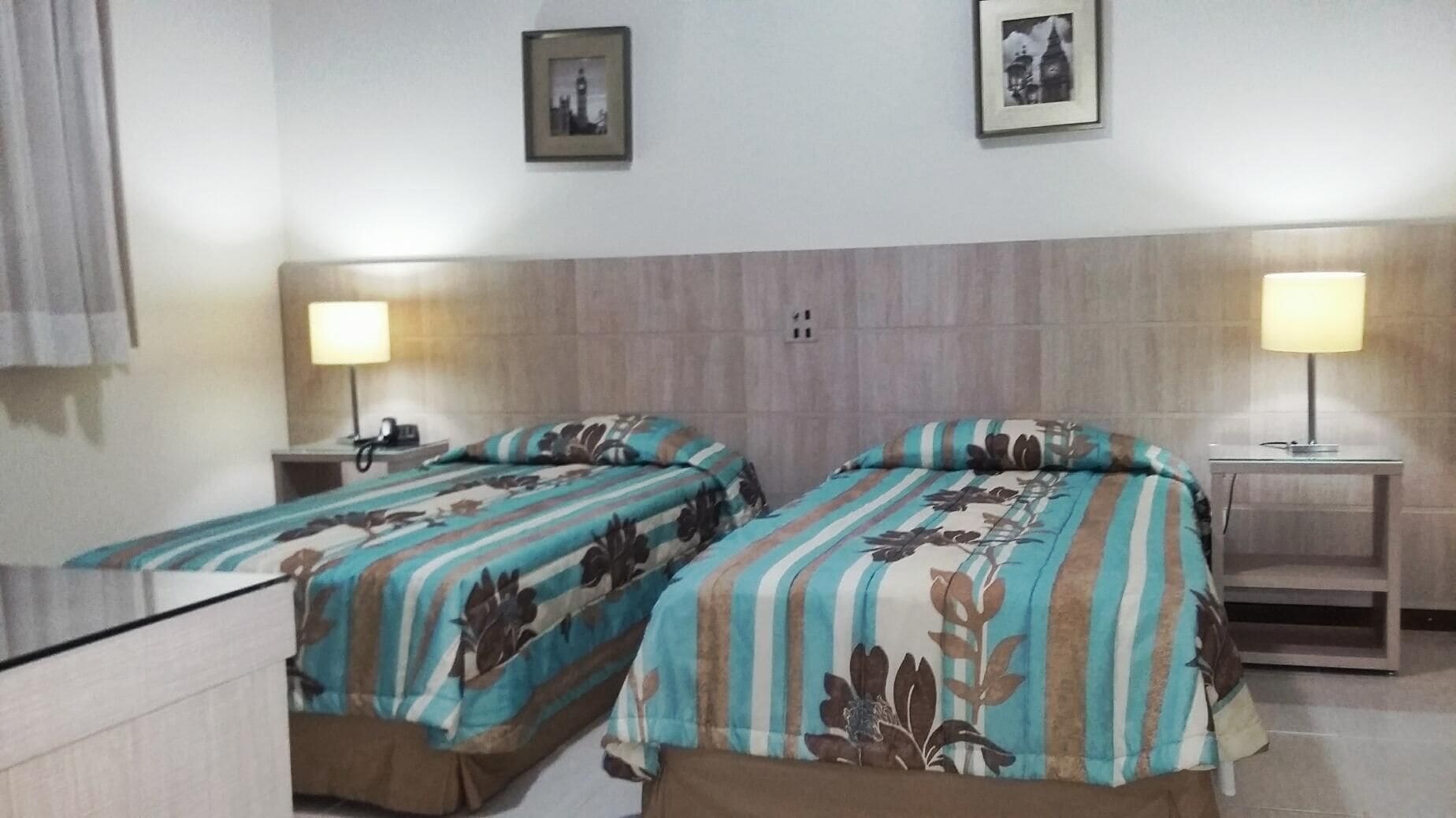 A hotel room with two single beds, each with a striped bedspread and a pillow. There are bedside tables with lamps, a wall-mounted power outlet, and two framed pictures above the beds.