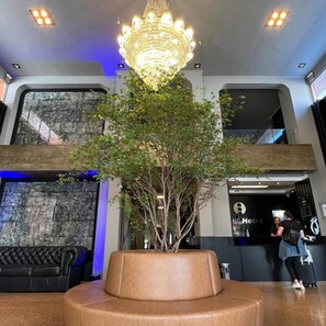 A lobby with a large, illuminated chandelier, a central planter with a tree, a black leather sofa, and a reception desk. - Arua Hotel (Presidente Prudente)