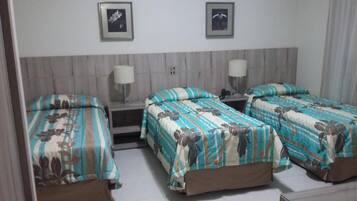 Quarto de hotel com três camas de solteiro, cada uma com colcha estampada com motivos florais, mesa de cabeceira com abajur e telefone fixado na parede.