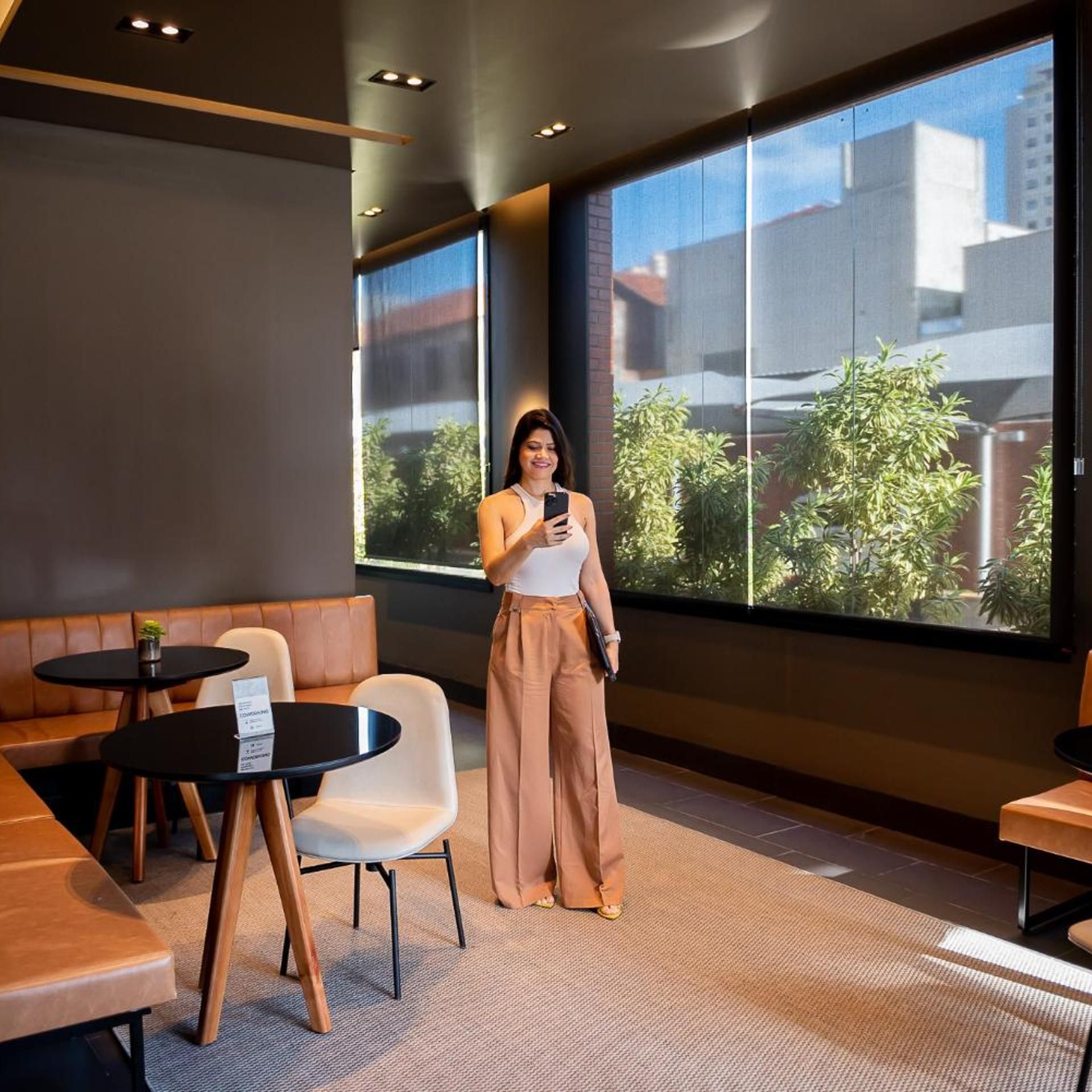 Um interior moderno com uma janela ampla, uma área de estar com mesa e cadeiras, e uma pessoa segurando um smartphone.