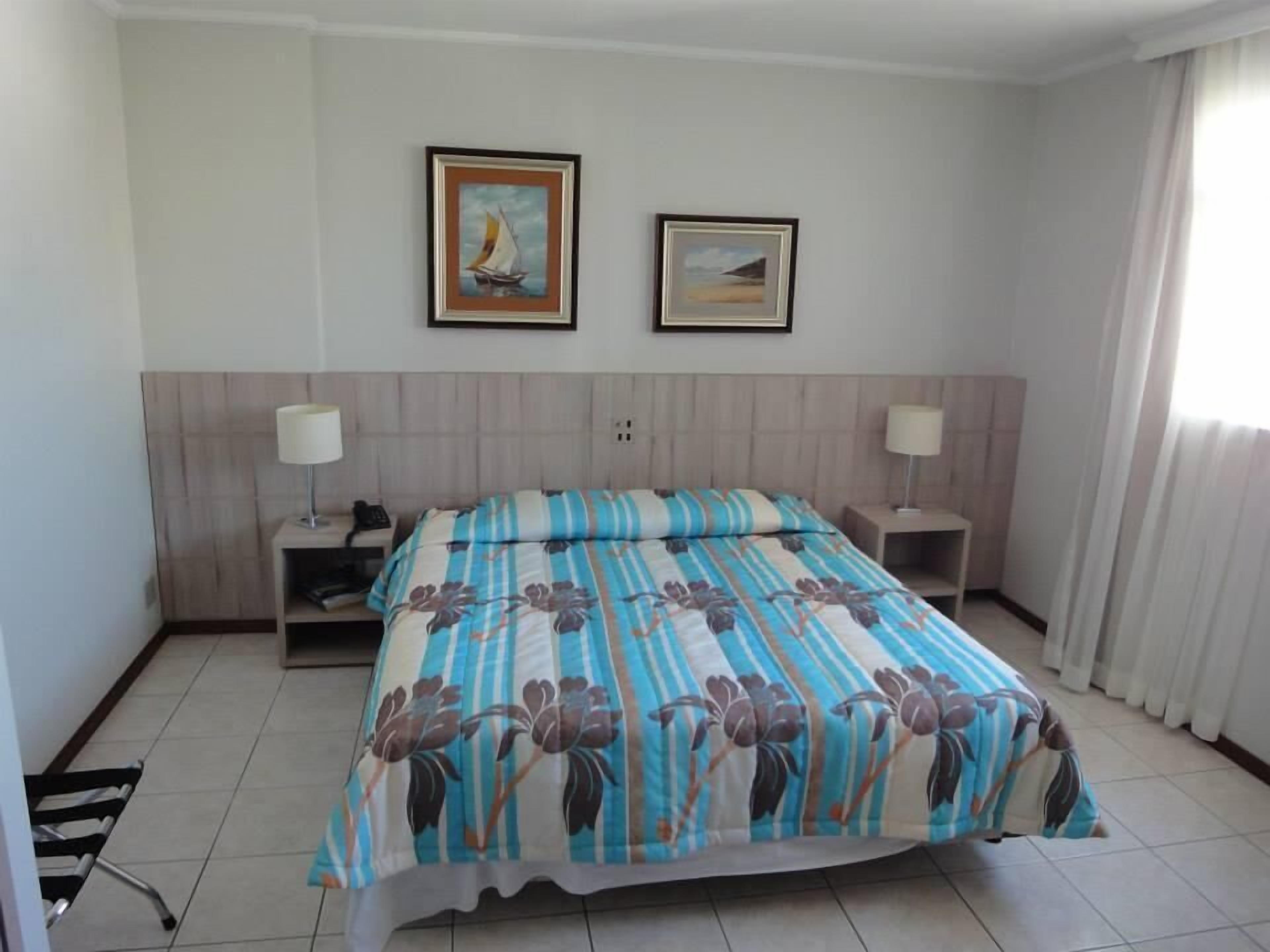 Quarto de hotel com uma cama, duas mesas de cabeceira, duas luminárias e duas imagens emolduradas na parede.