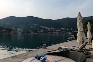 Vista al agua con sillones de descanso y sombrillas, un muelle y colinas con edificios al fondo.