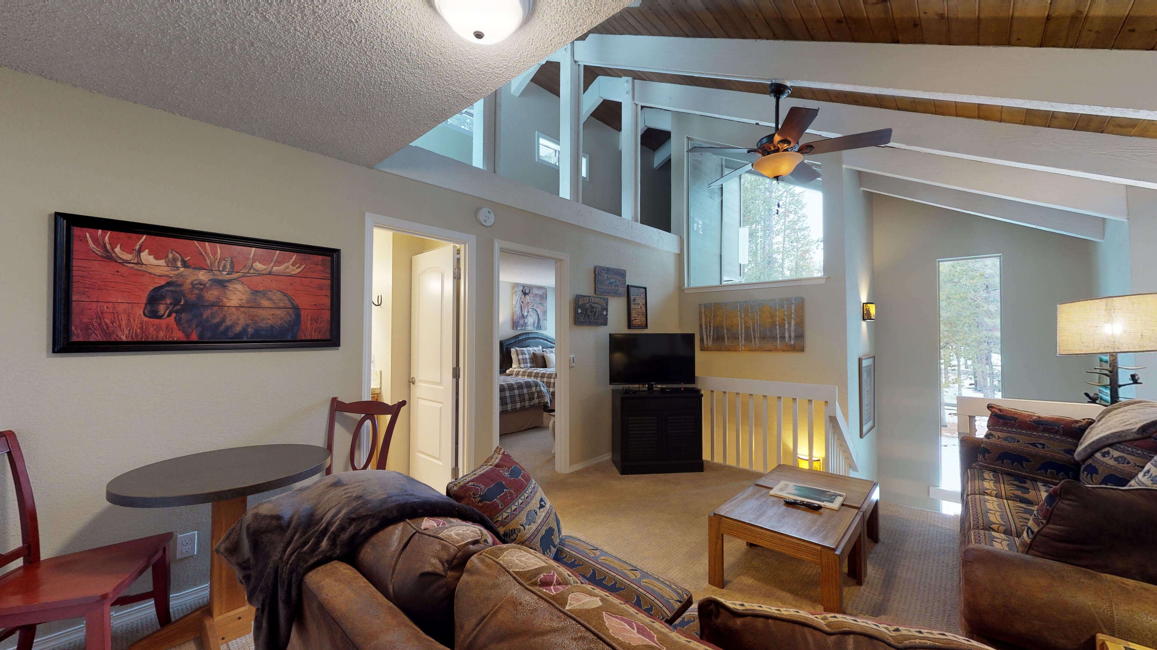 A spacious living room with a high ceiling, a ceiling fan, a mounted TV, and a large window offering a view of the outdoors.