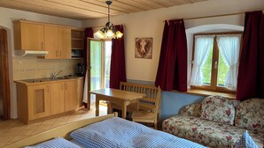 A cozy room with a wooden ceiling, a kitchenette, a dining table with chairs, a sofa, and a bed with checkered bedding. - Apartment 2 with balcony, up to 2 pers. (Rottau)