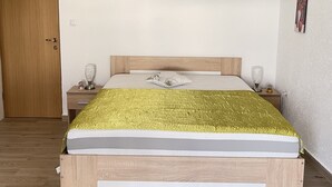 Un dormitorio con estructura de cama de madera, colchón blanco, manta texturizada dorada, mesita de noche con lámpara y un cuadro colgado en la pared.