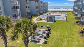 A pool area with lounge chairs, a barbecue grill, and a small building, surrounded by a well-maintained lawn and palm trees.
