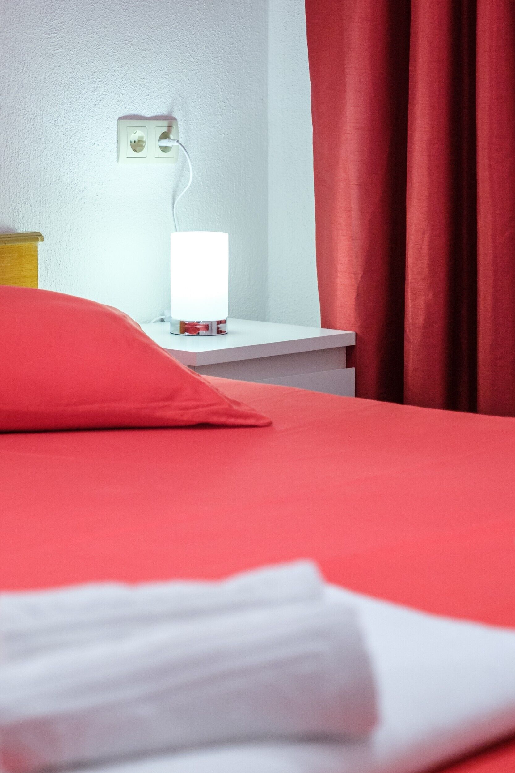 Una habitación de hotel con una cama, una mesita de noche con lámpara y un enchufe en la pared.