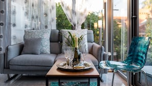 En moderne stue med en grå sofa, et træbord med en flaske og glas, og en blå lænestol ved et stort vindue.