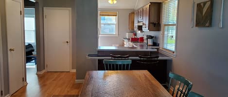 Uma cozinha com mesa e cadeiras, uma janela com persianas e uma porta que dá para outro cômodo.