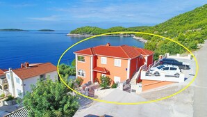 A coastal building with a red roof, adjacent to a road with parked cars, overlooking a body of water and green hills. - Holiday Home Adriatic View-Five Bedroom Holiday Home with Pool and Sea View (Blato)