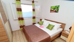Une chambre à coucher avec un lit, une table de chevet, une fenêtre avec des rideaux, un tableau accroché au mur et une vue sur la mer.