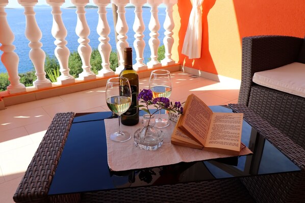 A balcony with a table set for two, featuring a bottle of wine, two wine glasses, a vase with flowers, and an open book.