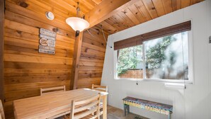 Interior de una cabaña de madera con una mesa de comedor, sillas, un banco y una ventana con persianas.