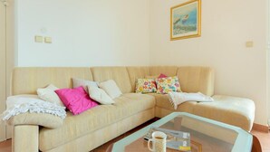 Un salon avec un canapé beige, des coussins colorés, une table basse en verre sur laquelle se trouvent un magazine et une tasse, et un tableau accroché au mur.