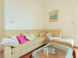 Ein Wohnzimmer mit einem beigefarbenen Sofa, bunten Kissen, einem Glastisch mit einer Zeitschrift und einer Tasse sowie einem Bild an der Wand.