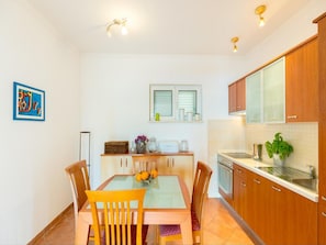A compact kitchen with wooden cabinets, a dining table with chairs, a window with blinds, and a small countertop with a sink and a potted plant.