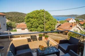 A balcony with wicker furniture, a glass table, and a view of a coastal town and hills. - Villa LA - Comfort One-Bedroom Apartment with Terrace and Sea View (Drvenik Veliki)