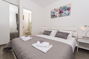 A neatly made bed with a grey quilt, two white towels, and purple flowers on the bed.